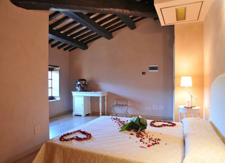 Camera Deluxe romantica con fiori e petali di rosa nel letto che formano due cuori luci accese e finestra nello sfondo travi di legno a vista nel soffitto