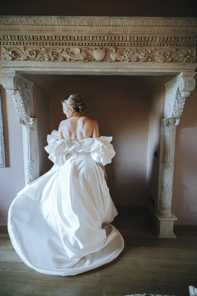 Sposa di spalle con abito moderno a sirena e scollo geometrico davanti a un grande camino antico scolpito in pietra.