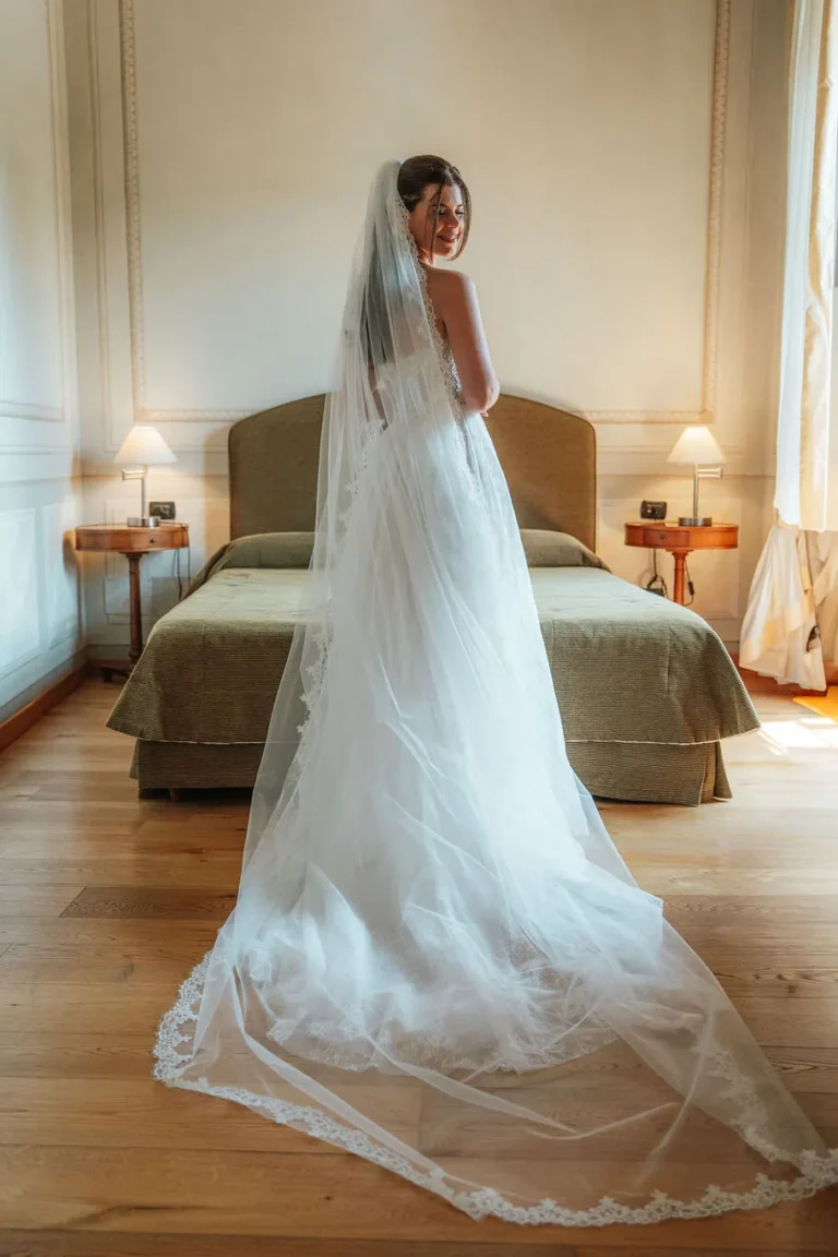 Sposa in piedi in una camera da letto d'epoca del Castello di Baccaresca con lungo velo ricamato in pizzo