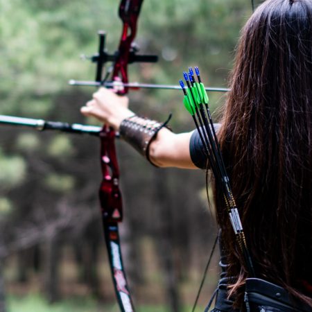 An athletic female aiming with bow and arrow towards the trees
